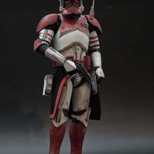 Può includere: Action figure dettagliata di un clone trooper in un'armatura rossa, bianca e nera. La figura tiene in mano una pistola blaster e ha un elmetto con segni rossi. La figura è in uno studio.