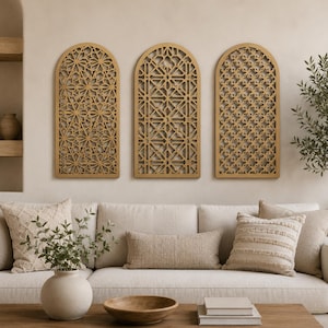 Op de afbeelding: Drie gouden, boogvormige wandpanelen met ingewikkelde geometrische uitgesneden ontwerpen. Ze zijn gemonteerd boven een beige bank met decoratieve kussens en een houten salontafel. De kamer heeft een neutraal kleurenpalet.