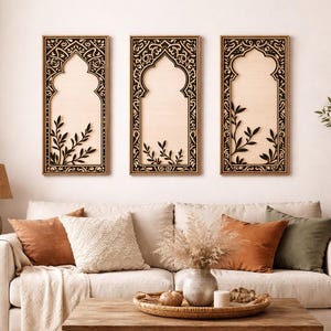Op de afbeelding: Drie rechthoekige houten wandkunstwerken met een boogvormig ontwerp. Elk stuk heeft een lichtgekleurde achtergrond met een zwarte, sierlijke rand en een zwart botanisch ontwerp. De kunst wordt tentoongesteld boven een crèmekleurige bank met decoratieve kussens.