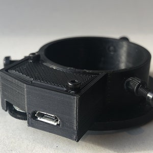 May include: A black 3D printed camera mount with a micro USB port. The mount has a threaded hole on the bottom and a screw on the side.