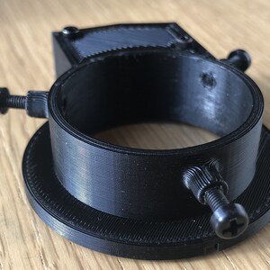May include: A black 3D printed camera lens adapter with two adjustable screws. The adapter has a circular opening and a flat base.