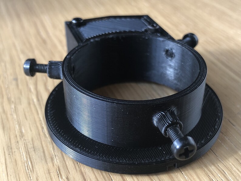 May include: A black 3D printed camera lens adapter with two adjustable screws. The adapter has a circular opening and a flat base.