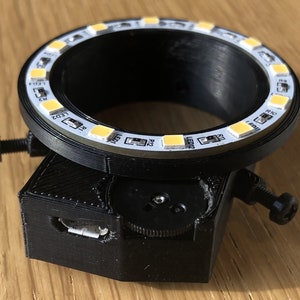 May include: A black 3D printed ring light with a USB port and a knob for adjusting the light. The ring light has 12 white LED lights around the circumference.