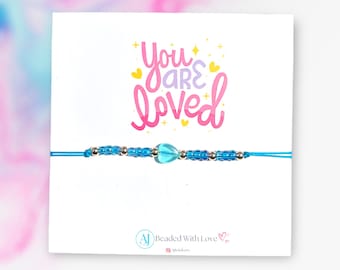 May include: A blue beaded bracelet with a light blue heart charm. The bracelet has the words "You are loved" written on a white card behind it.