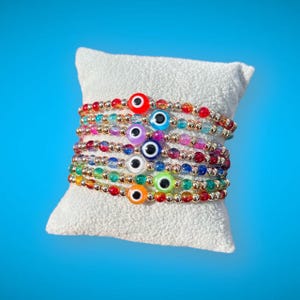 Puede incluir: Una pila de pulseras de cuentas de colores con dijes de ojo turco. Cada pulsera presenta un color de cuentas diferente y un dije de ojo turco de color diferente.