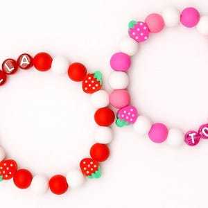 Peut inclure: Deux bracelets de perles. L'un est rouge, blanc et sur le thème de la fraise avec le nom "BELLA" épelé. L'autre est rose, blanc et sur le thème de la fraise avec le nom "TOBY" épelé. Les bracelets sont sur fond blanc.
