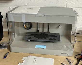 May include: A gray 3D printer with a clear plastic enclosure. The printer is on a wooden surface and has a black build plate with a printed object on it. The printer has a digital display and a Markforged logo on the front.