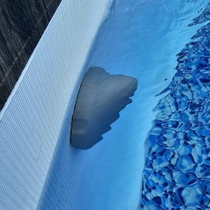 Op de afbeelding: Een close-up van een grijze zwembadskimmer, gedeeltelijk ondergedompeld in een blauw zwembad met een witte betegelde rand.