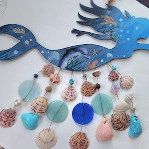 May include: A blue wooden mermaid wall hanging featuring painted sea life including a dolphin, turtle, and octopus. Dangling from the mermaid are multiple strands of assorted seashells, blue and clear glass discs, and small decorative beads.