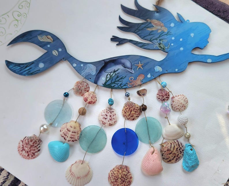 May include: A blue wooden mermaid wall hanging featuring painted sea life including a dolphin, turtle, and octopus. Dangling from the mermaid are multiple strands of assorted seashells, blue and clear glass discs, and small decorative beads.