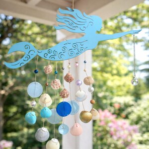 May include: A light blue metal mermaid silhouette hanging mobile featuring intricate cut-out details. Strands of twine hang from the mermaid, decorated with various seashells, blue and clear glass discs, and beads, ending with a crystal prism.