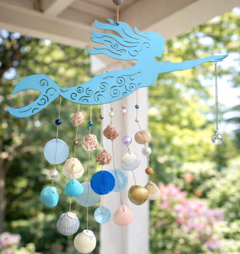 May include: A light blue metal mermaid silhouette hanging mobile featuring intricate cut-out details. Strands of twine hang from the mermaid, decorated with various seashells, blue and clear glass discs, and beads, ending with a crystal prism.