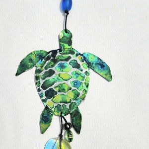 May include: A green and blue painted turtle wind chime with a heart, beads, and a bell. The turtle is made of metal and has a black cord.