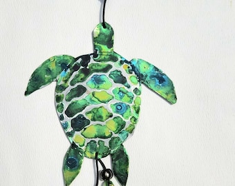 May include: A green and blue painted turtle wind chime with a heart, beads, and a bell. The turtle is made of metal and has a black cord.