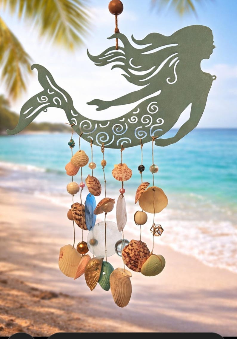 May include: A sage green metal mermaid silhouette hanging mobile featuring intricate swirl cutouts. Multiple strands of twine hang from the mermaid, decorated with various seashells, smooth wooden beads, clear glass spheres, and blue glass accents.