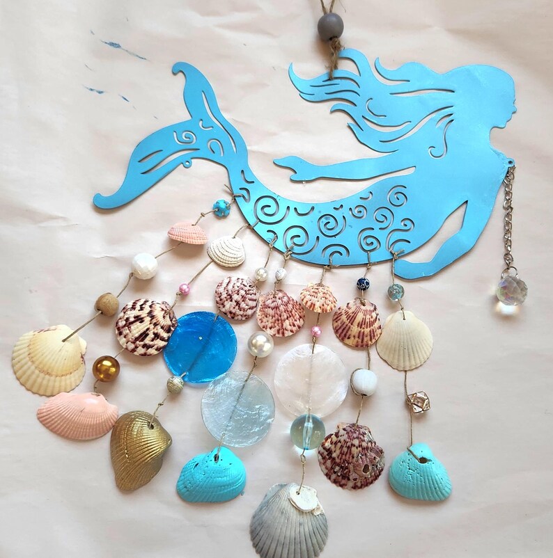 May include: A light blue metal mermaid silhouette wall hanging featuring intricate scrollwork cutouts. Multiple strands of twine hang from the mermaid, decorated with various seashells, small glass beads, and a clear crystal pendant.