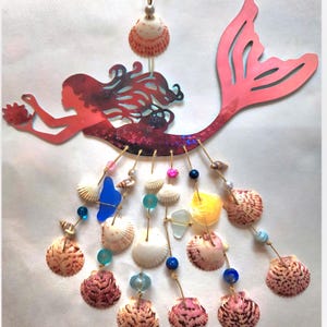 May include: A hanging wall decoration featuring a red mermaid silhouette with a long tail. Below the mermaid, multiple strands of twine are adorned with various seashells, blue sea glass, and colorful beads in shades of pink, blue, and yellow.