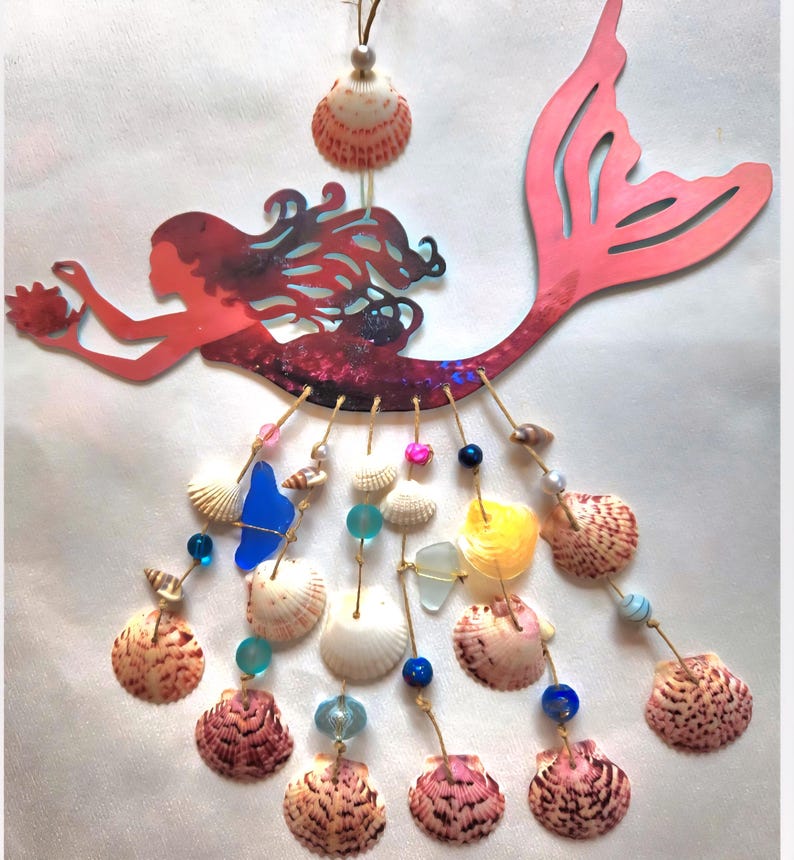 May include: A hanging wall decoration featuring a red mermaid silhouette with a long tail. Below the mermaid, multiple strands of twine are adorned with various seashells, blue sea glass, and colorful beads in shades of pink, blue, and yellow.
