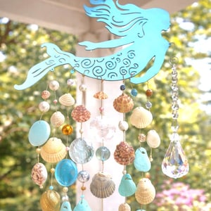 May include: A hanging mobile featuring a light blue metal mermaid silhouette with a swirl pattern on the tail. Strands of twine hang from the mermaid, decorated with various seashells, clear glass beads, blue glass stones, and a large crystal pendant.