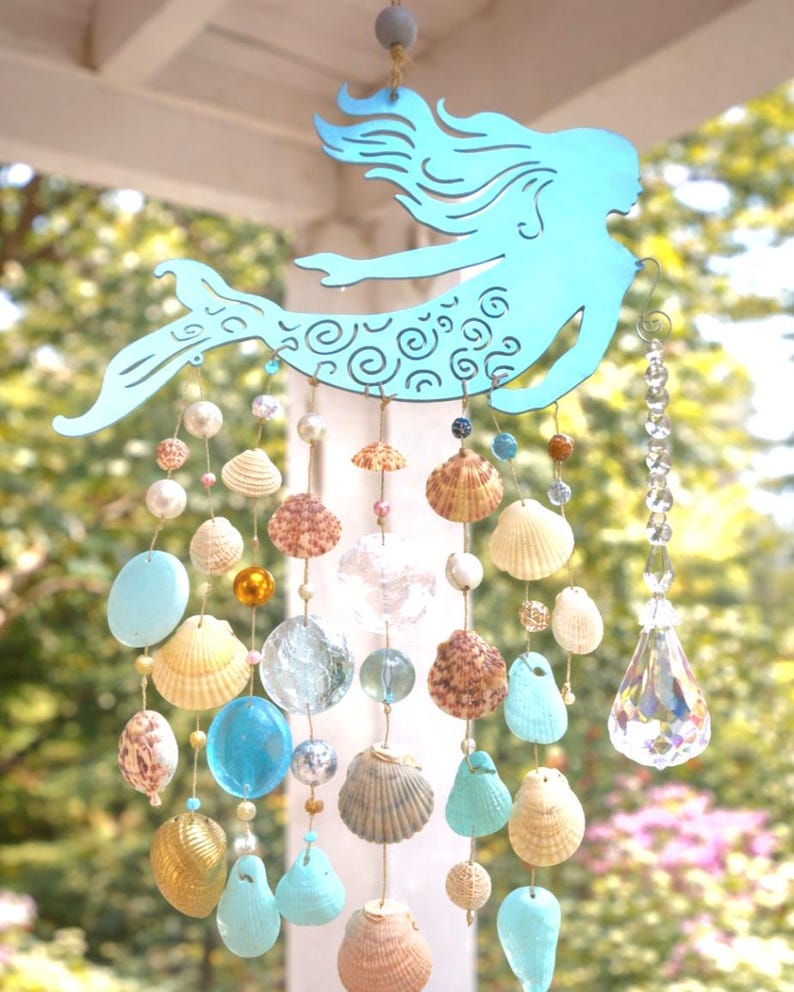 May include: A hanging mobile featuring a light blue metal mermaid silhouette with a swirl pattern on the tail. Strands of twine hang from the mermaid, decorated with various seashells, clear glass beads, blue glass stones, and a large crystal pendant.