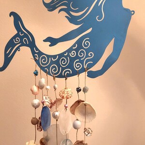 May include: A blue metal mermaid silhouette wall hanging featuring intricate white swirl patterns on the tail. Multiple strands of jute twine hang from the mermaid, decorated with various seashells, white pearls, and translucent glass beads.