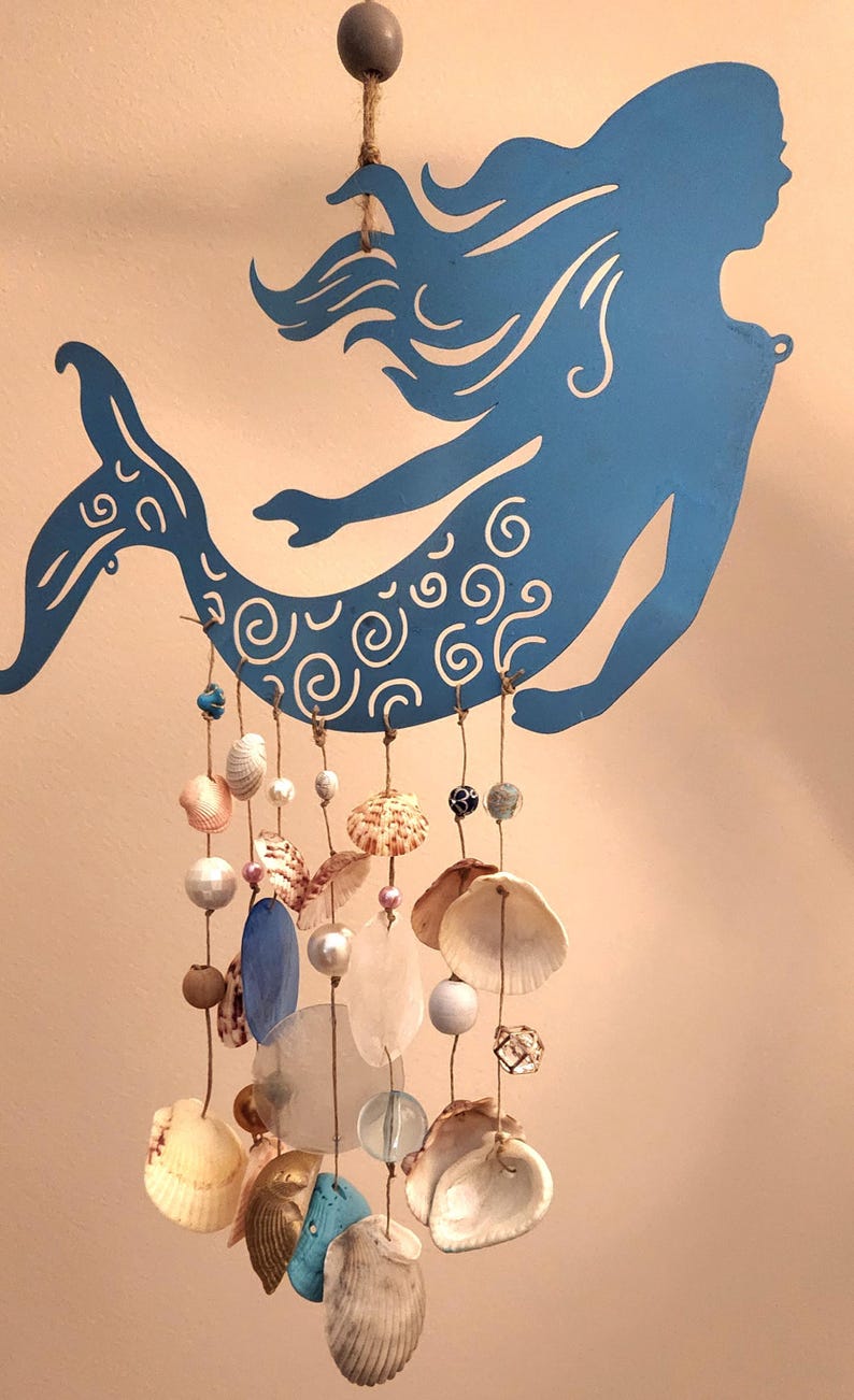 May include: A blue metal mermaid silhouette wall hanging featuring intricate white swirl patterns on the tail. Multiple strands of jute twine hang from the mermaid, decorated with various seashells, white pearls, and translucent glass beads.