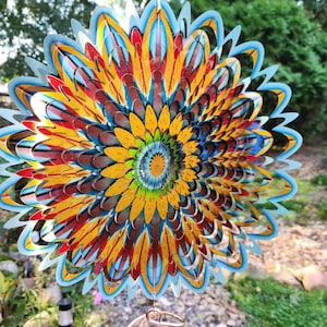 May include: A circular metal wind spinner featuring a multi-layered, flower-like design with pointed petals. The color palette includes vibrant shades of yellow, orange, red, green, and blue, arranged in a radial pattern that creates a visual depth.