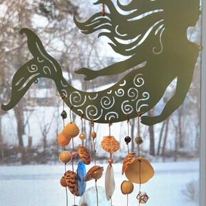 May include: A sage green metal mermaid silhouette hanging ornament featuring intricate swirl cutouts. Suspended from the mermaid are multiple strands adorned with various seashells, translucent blue glass discs, and small metallic beads.