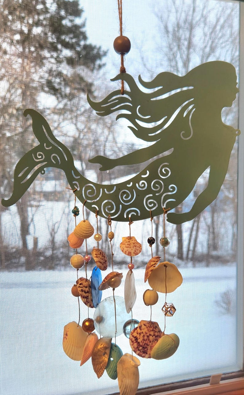 May include: A sage green metal mermaid silhouette hanging ornament featuring intricate swirl cutouts. Suspended from the mermaid are multiple strands adorned with various seashells, translucent blue glass discs, and small metallic beads.