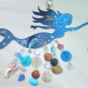 May include: A blue wooden mermaid wall hanging featuring illustrations of a dolphin, sea turtle, crab, and octopus. Dangling from the mermaid are strings of various seashells, blue glass discs, white beads, and a clear crystal prism pendant.