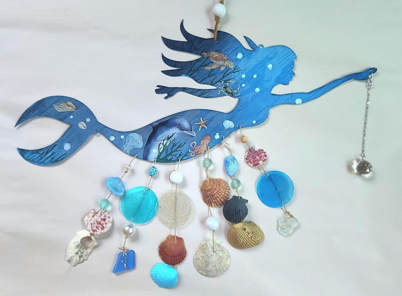 May include: A blue wooden mermaid wall hanging featuring illustrations of a dolphin, sea turtle, crab, and octopus. Dangling from the mermaid are strings of various seashells, blue glass discs, white beads, and a clear crystal prism pendant.