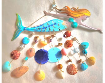 May include: A wall hanging featuring a metal mermaid with long orange hair and a blue tail. Suspended from the mermaid are multiple strings adorned with various seashells, colorful beads, and two circular blue discs in the center.