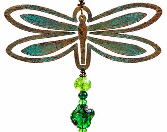 May include: A hanging ornament featuring a copper-toned metal dragonfly with a textured teal patina finish. The dragonfly is accented with a vertical arrangement of green glass beads and a copper-toned metal bell at the bottom.