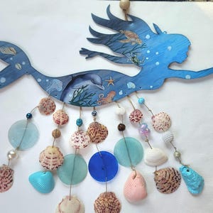 May include: A blue wooden mermaid wall hanging featuring painted ocean life including a dolphin, turtle, and octopus. Dangling from the mermaid are multiple strands of natural seashells, blue and clear glass discs, and beads, ending with a crystal.