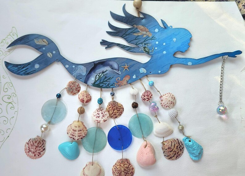 May include: A blue wooden mermaid wall hanging featuring painted ocean life including a dolphin, turtle, and octopus. Dangling from the mermaid are multiple strands of natural seashells, blue and clear glass discs, and beads, ending with a crystal.