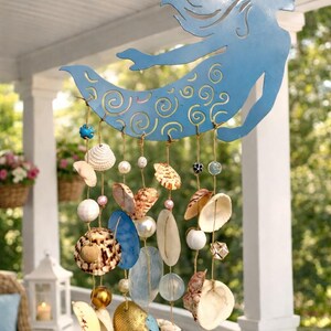 May include: A hanging wind chime featuring a light blue metal mermaid silhouette with cut-out swirl patterns. Strands of jute twine hang from the mermaid, decorated with various seashells, white and gold beads, and blue glass accents.