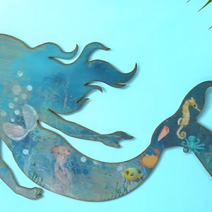 May include: A flat, cutout wooden wall decoration shaped like a mermaid, featuring a blue-toned finish with painted illustrations of a seahorse, an octopus, a crab, a fish, a jellyfish, and a seashell, with a small metal hanging loop attached.