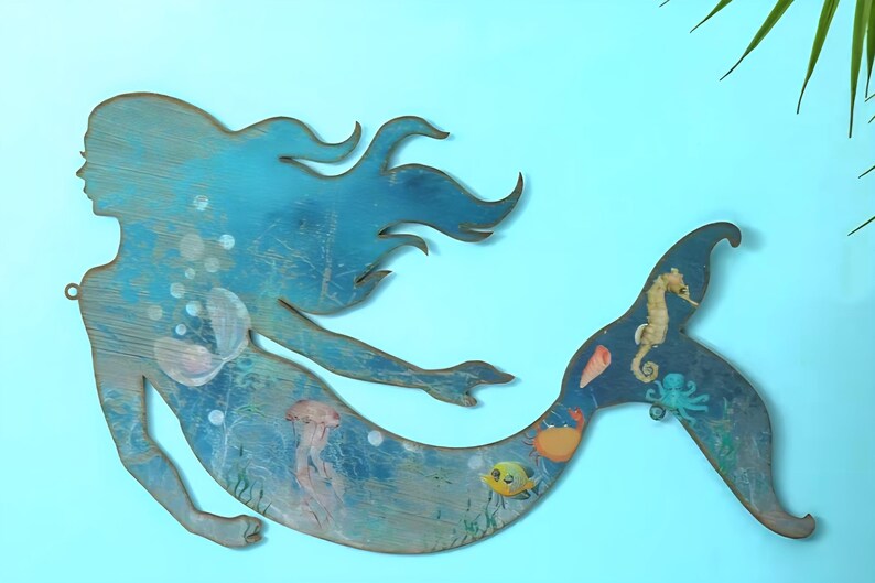 May include: A flat, cutout wooden wall decoration shaped like a mermaid, featuring a blue-toned finish with painted illustrations of a seahorse, an octopus, a crab, a fish, a jellyfish, and a seashell, with a small metal hanging loop attached.