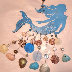 May include: A light blue metal mermaid silhouette wall hanging featuring intricate swirl cutouts. Dangling from the mermaid are multiple strands of twine adorned with various seashells, including brown, white, pink, and gold-painted shells, along with blue, white, and clear beads.