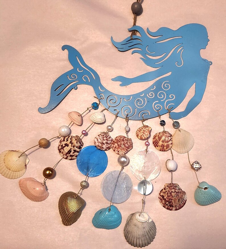 May include: A light blue metal mermaid silhouette wall hanging featuring intricate swirl cutouts. Dangling from the mermaid are multiple strands of twine adorned with various seashells, including brown, white, pink, and gold-painted shells, along with blue, white, and clear beads.