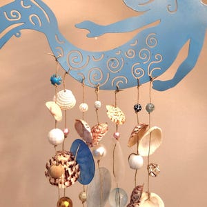 May include: A light blue metal mermaid silhouette serves as a hanger for a mobile featuring multiple strands of twine. These strands are adorned with various seashells, including white, brown, and gold-toned varieties, interspersed with glass and plastic beads.