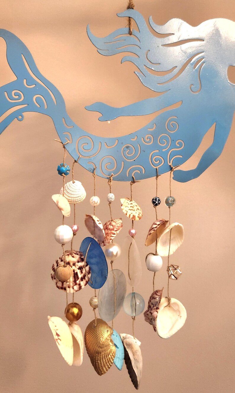 May include: A light blue metal mermaid silhouette serves as a hanger for a mobile featuring multiple strands of twine. These strands are adorned with various seashells, including white, brown, and gold-toned varieties, interspersed with glass and plastic beads.