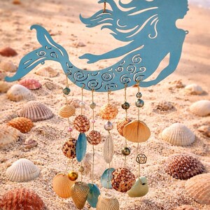 May include: A light blue metal mermaid silhouette hanging ornament featuring intricate swirl cutouts. Suspended from the mermaid are multiple gold-toned wires adorned with various natural seashells, small pearls, and metallic beads in shades of tan, brown, and cream.