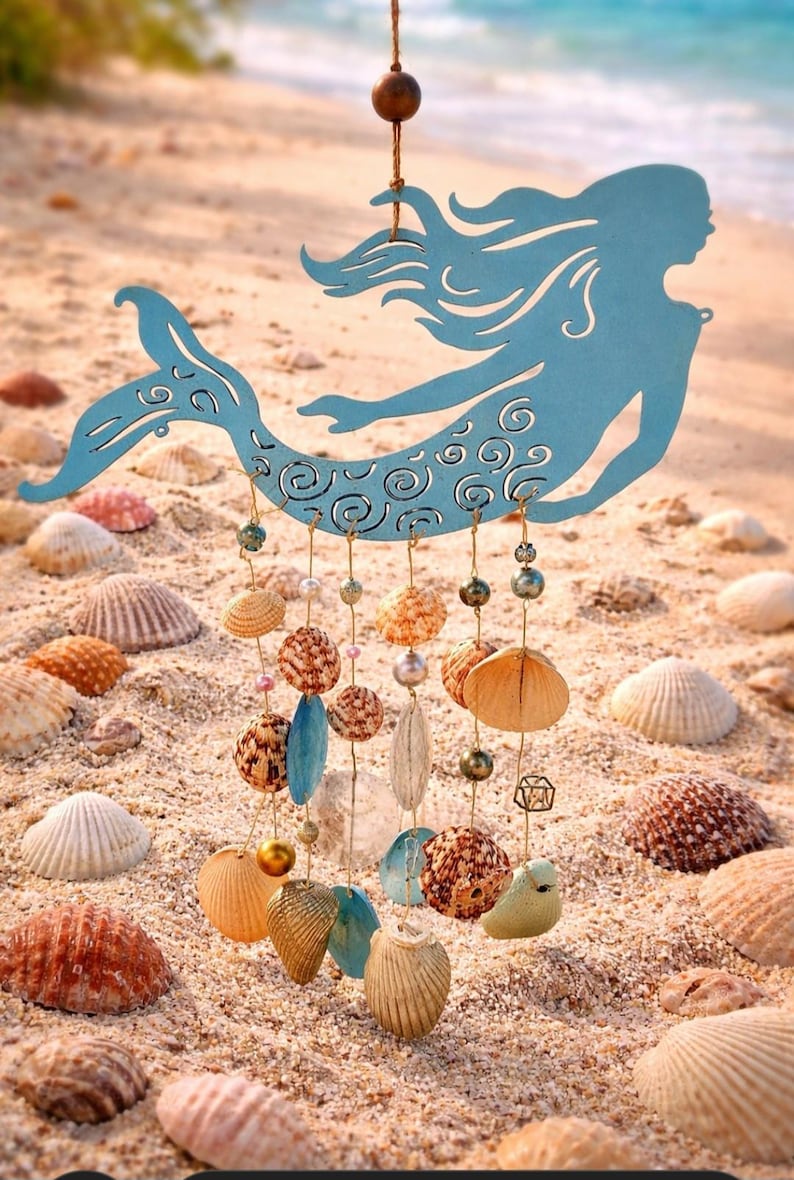 May include: A light blue metal mermaid silhouette hanging ornament featuring intricate swirl cutouts. Suspended from the mermaid are multiple gold-toned wires adorned with various natural seashells, small pearls, and metallic beads in shades of tan, brown, and cream.