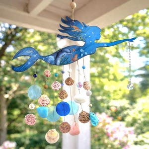 May include: A hanging mobile featuring a blue mermaid silhouette painted with sea life including a dolphin, jellyfish, and starfish. Strands of natural seashells, blue glass discs, and small beads hang from the mermaid, ending with a crystal prism.