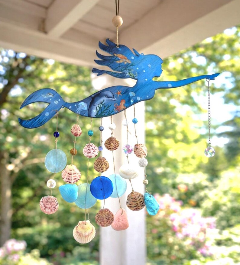 May include: A hanging mobile featuring a blue mermaid silhouette painted with sea life including a dolphin, jellyfish, and starfish. Strands of natural seashells, blue glass discs, and small beads hang from the mermaid, ending with a crystal prism.