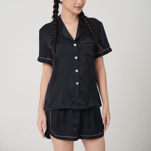 May include: A black satin pajama set with short sleeves and shorts. The top has a collared neckline and button closure. The shorts have a pink trim.