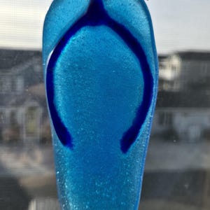May include: A blue glass flip-flop ornament with a dark blue thong design. The ornament is suspended by a thin gold-colored wire. The glass has a textured, sparkling appearance. The background shows a blurred view of buildings and a window.