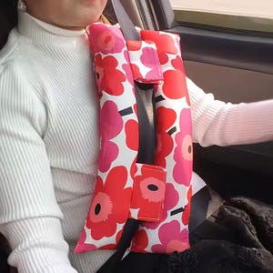 May include: A white, red, and pink floral patterned car seat belt cushion. The cushion has two black straps and a black buckle.
