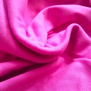 May include: Close-up of a vibrant fuchsia fabric with soft folds and subtle textures. The material appears to be a knit or jersey, with a smooth, slightly shiny surface. The image highlights the fabric's color and drape.